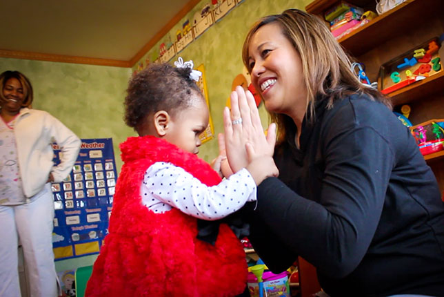 Cute toddler girl clapping with specialist as mom looks on in the background image.