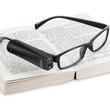 Orcam is seen connected to the side of a glasses frame. The glasses sit on top of a book.