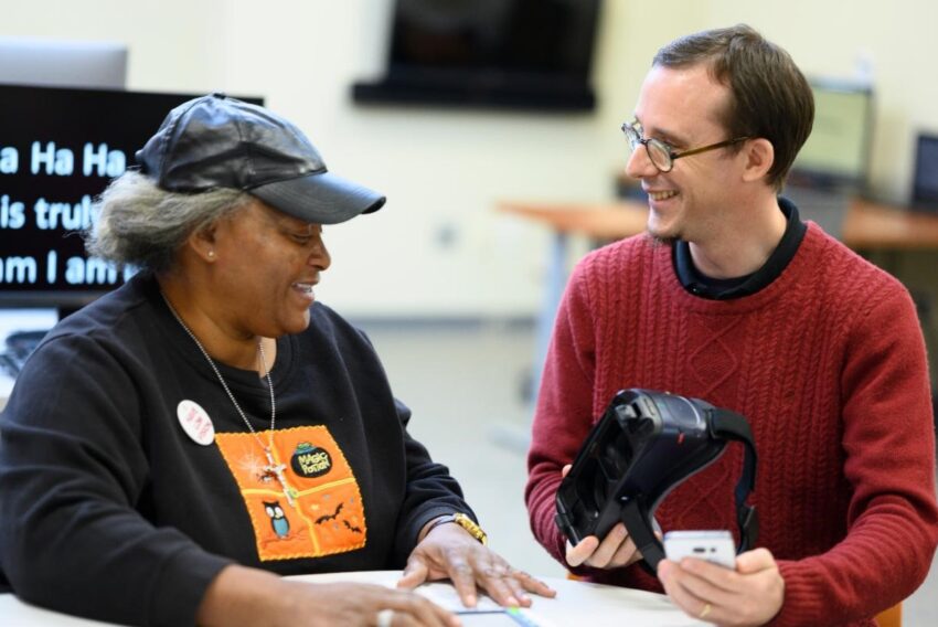 A woman sits to the left of a man, both of who are smiling. Then man is holding a wearable electronic magnification device, and seems to be demonstrating it to the woman.