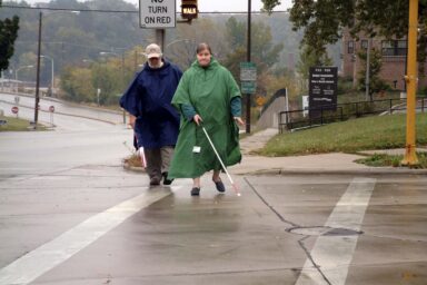 Two people crossing in a crosswalk in rainy weather, using white-tipped cane