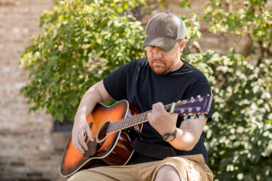 Chris is strumming his guitar outside on a summer day.