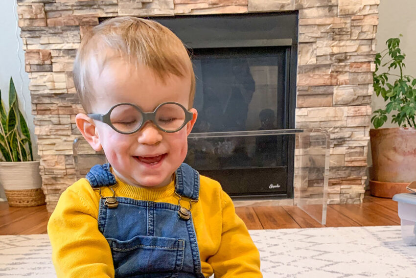 Toddler boy in overalls, wearing glasses, smiling as he strums a ukelele.