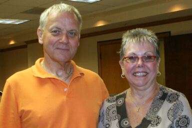 A man in an orange shirt is standing with his wife, in an abstract-patterned dress. Both are smiling