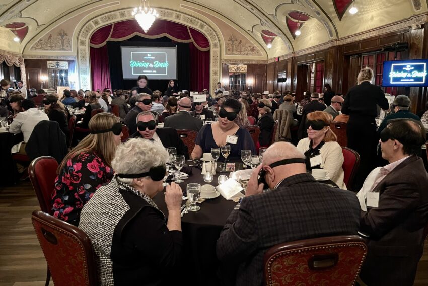 Dozens of attendees seated at tables for 8, wearing blindfolds while eating dinner.