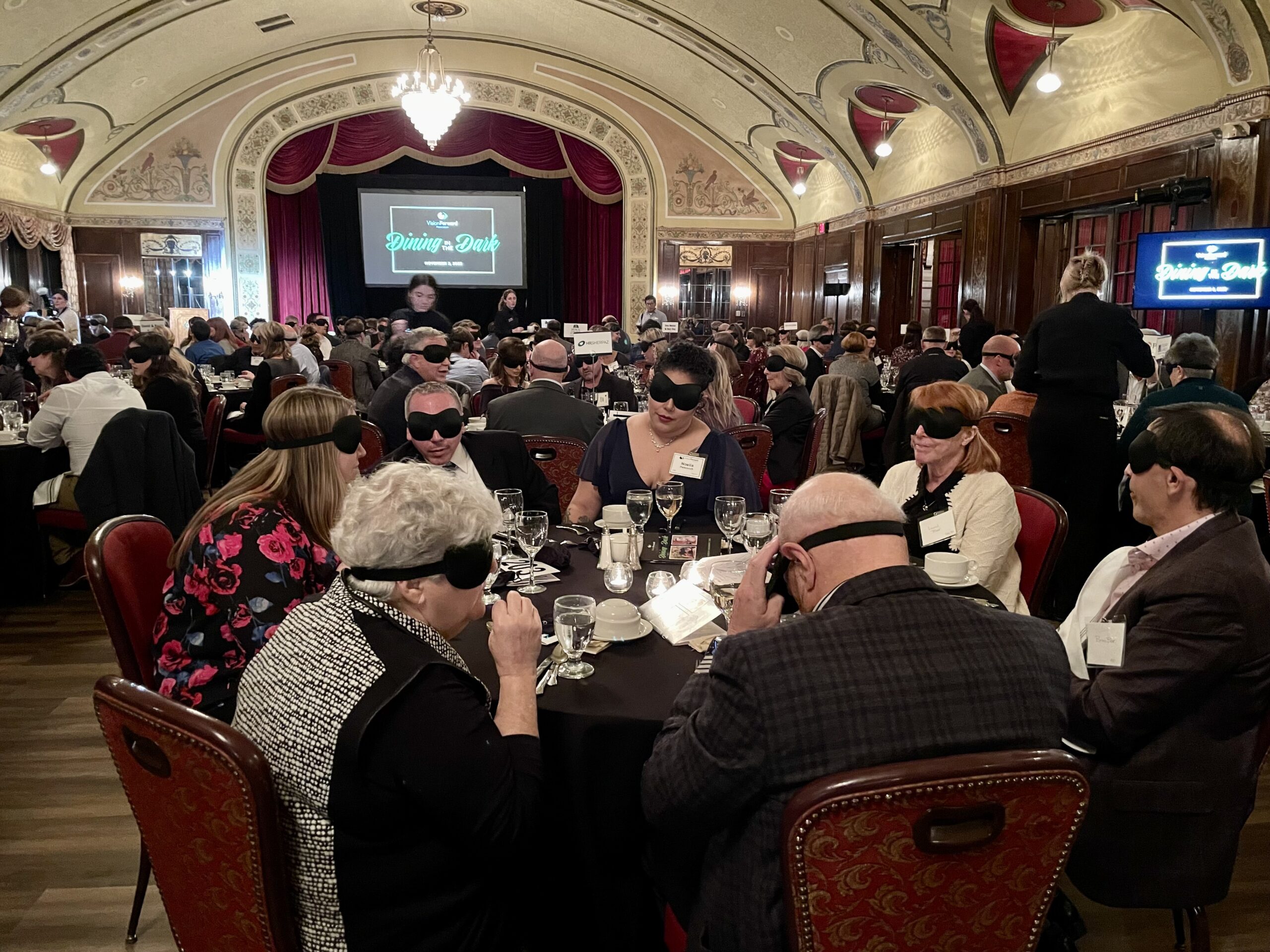 Dozens of attendees seated at tables for 8, wearing blindfolds while eating dinner.