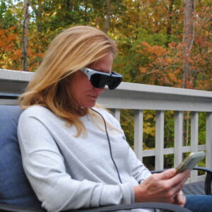 A woman sits outdoors. She wears the eyedaptic 5, using it to look at her smartphone.