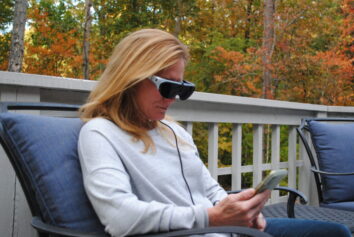 A woman sits outdoors. She wears the eyedaptic 5, using it to look at her smartphone.