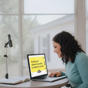 A woman looks at a computer screen, where magnified text is shown against a yellow background. Next to the computer the Orcam Read 3. sist in its stand.