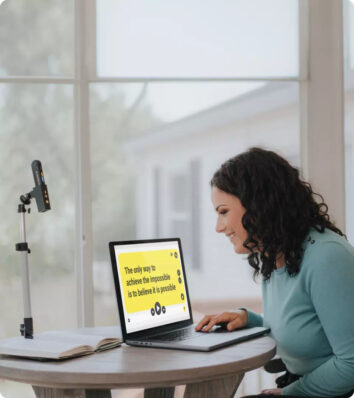 A woman looks at a computer screen, where magnified text is shown against a yellow background. Next to the computer the Orcam Read 3. sist in its stand.