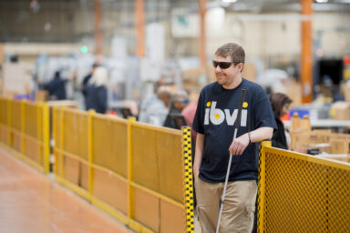 Chris leaning against a protective barrier with his ibvi tshirt with dark glasses on and his cane