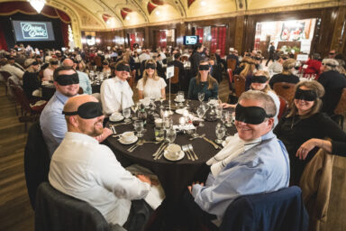 people dressed in formal attire seated around tables with black eye masks on