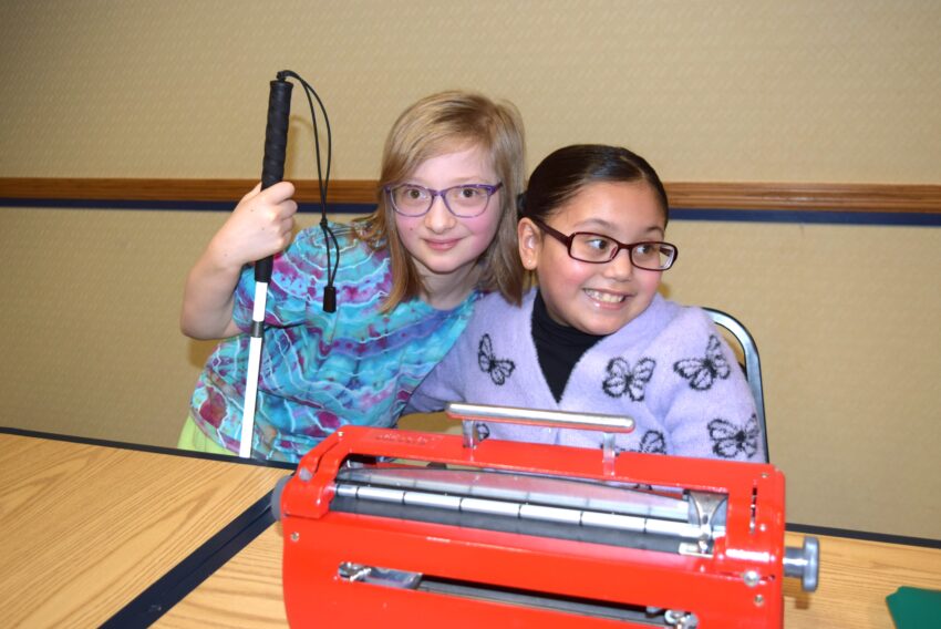 2 school aged visually impaired girls smiling behind a brailler