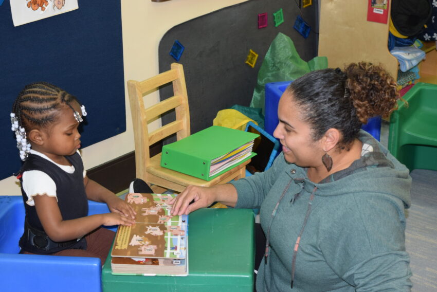 visually impaired toddler with book open and teacher helping her read braille