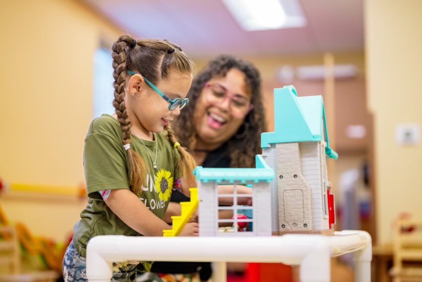 visually impaired young girl plays with dollhouse while teacher smiles