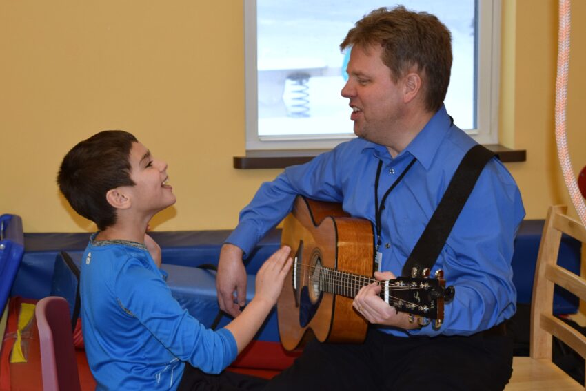 Little boy in blue shirt singing and helping teacher strum the guitar while he's playing it