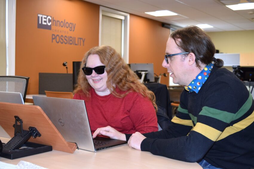 young woman on computer with dark glasses on being helped by man with glasses.