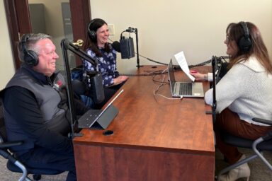 3 people sitting together with headphones on smiling and talking into microphones