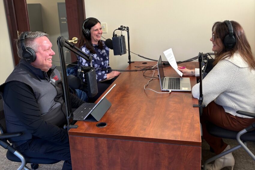 3 people sitting together with headphones on smiling and talking into microphones