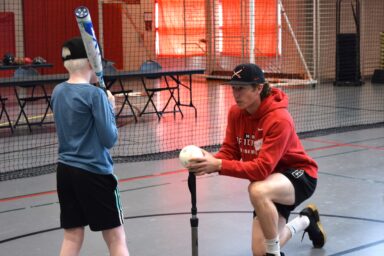 College age boy with baseball cap on kneeling with his hands around a baseball placed on a stand. Little boy with baseball bat waiting to hit the ball.