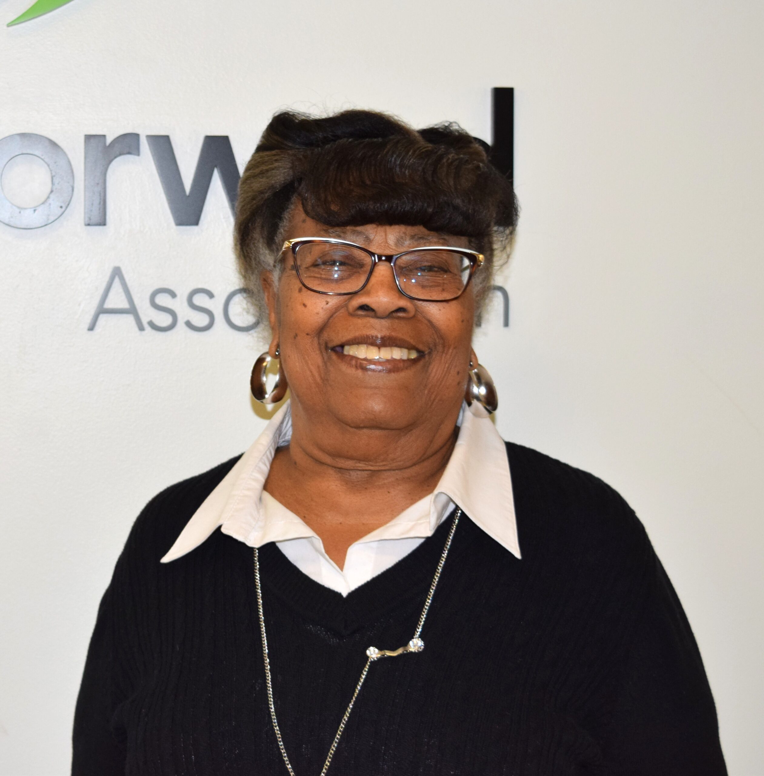 Older black woman dressed in black sweater and white blouse with glasses and hoop earrings on.