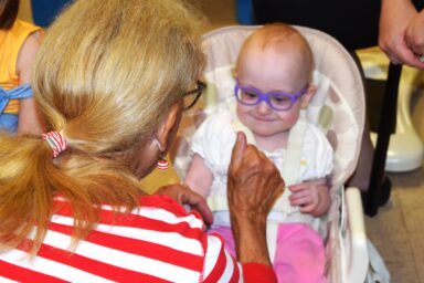 Baby with glasses smiling while teacher holds up hands