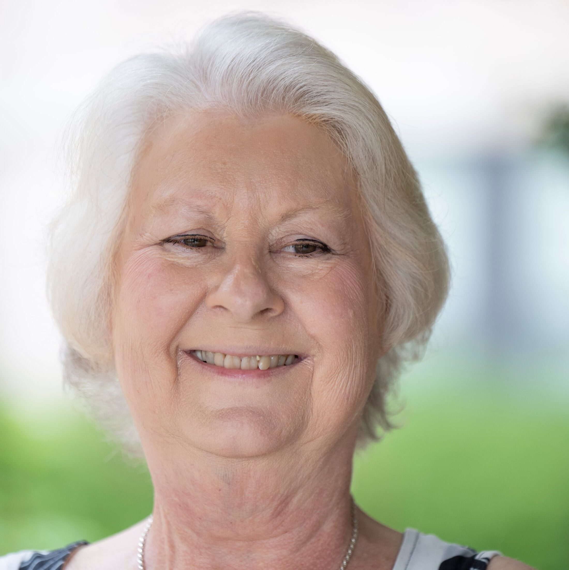 older woman with white shorter hair and a big smile standing outdoors in the summer.