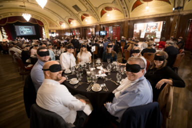 A nicely dressed group of people seated at a table in a fancy room with blindfolds on. There are meals in front of them. They're all smiling and turning toward the camera.