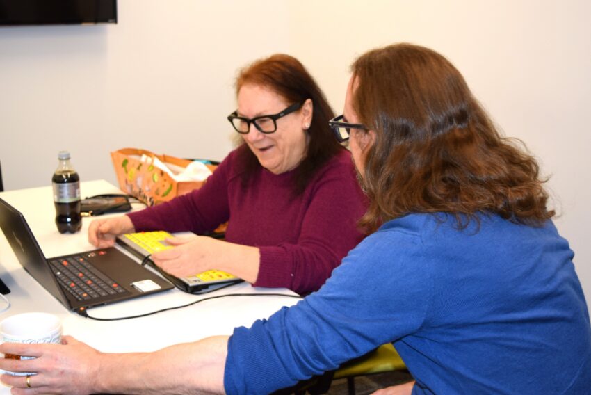 Dark haired older woman with glasses is working on her laptop computer and a man is assisting her with instructions while she works