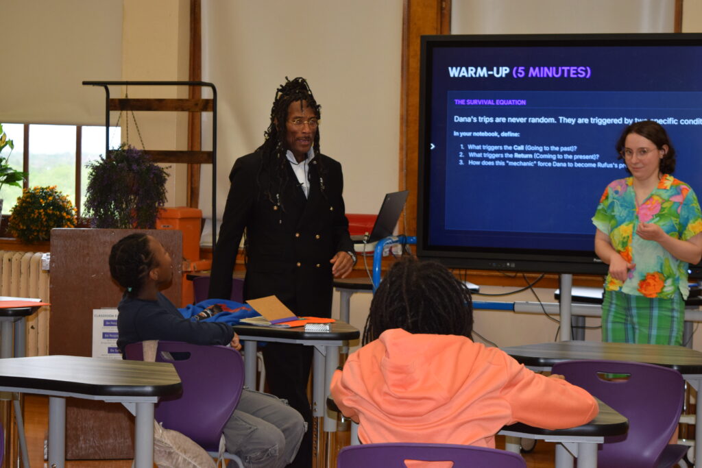 A man is standing in front of a classroom full of children with a tv behind him showing a presentation